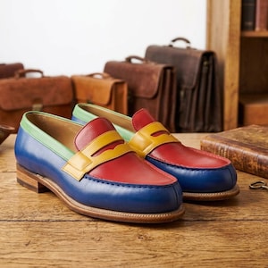 Puede incluir: Un par de mocasines de cuero multicolores. Los zapatos tienen puntera azul, empeine rojo, correa amarilla y ribete verde. Se exhiben sobre una superficie de madera, con maletines de cuero en el fondo. Estilo clásico.