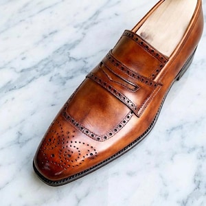 May include: A tan leather penny loafer shoe featuring a burnished finish, a decorative strap across the vamp, and intricate brogue detailing with perforated patterns along the toe cap and the edges of the saddle strap.