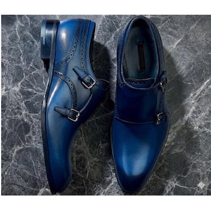 May include: A pair of blue leather double monk strap shoes. The shoes have a glossy finish, decorative perforations, and silver-colored buckles. The shoes are displayed on a gray marble surface.