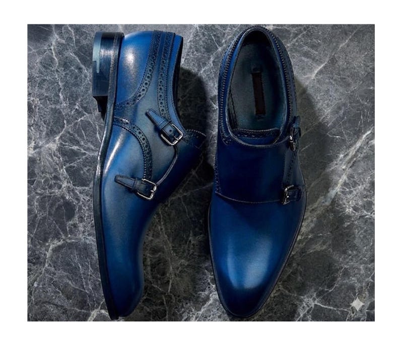 May include: A pair of blue leather double monk strap shoes. The shoes have a glossy finish, decorative perforations, and silver-colored buckles. The shoes are displayed on a gray marble surface.