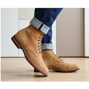 May include: Tan suede boots with a cap toe and lace-up design. The boots have a brown sole and are worn with blue jeans with a turned-up cuff. The boots are on a light wood floor.