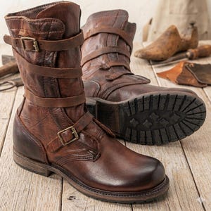 Puede incluir: Botas de cuero marrón con múltiples correas y cierres de hebilla. Las botas tienen una apariencia robusta y desgastada y una suela gruesa. Las botas se exhiben sobre una superficie de madera, lo que sugiere un estilo artesanal o vintage. Las botas son de media caña.