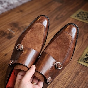 May include: A pair of brown leather double monk strap shoes featuring a textured suede vamp panel, polished silver-tone metal buckles, and a burnished finish on the leather toe and heel sections, showcasing a classic silhouette with dual straps.