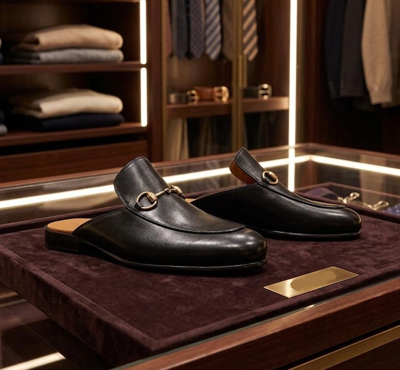 Puede incluir: Un par de mules de cuero negro con detalles met&aacute;licos dorados. Los zapatos se exhiben sobre una superficie de terciopelo marr&oacute;n oscuro, mostrando su dise&ntilde;o elegante y artesan&iacute;a. Las mules tienen una est&eacute;tica cl&aacute;sica y refinada.
