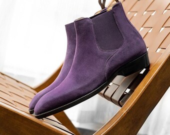 May include: A pair of purple suede Chelsea boots with black soles and elastic side panels. The boots are displayed on a wooden chair with a woven seat. The boots have a pull tab at the back.