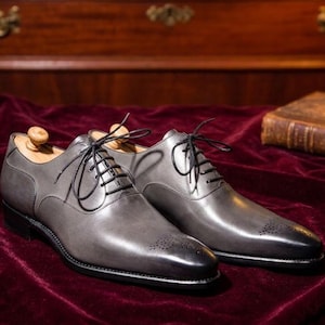 May include: A pair of grey leather dress shoes with black laces and an Oxford design. The shoes have a polished finish and are displayed on a burgundy velvet surface.