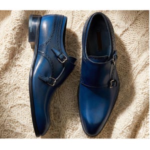 May include: A pair of blue leather dress shoes with double monk straps and silver buckles. The shoes have a polished finish and are set against a textured, off-white background.