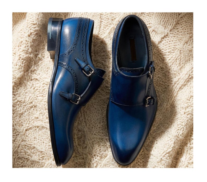 May include: A pair of blue leather dress shoes with double monk straps and silver buckles. The shoes have a polished finish and are set against a textured, off-white background.