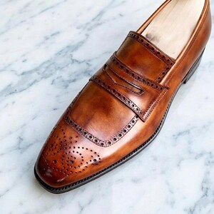 May include: A tan leather penny loafer shoe featuring a burnished finish, a decorative strap across the vamp, and intricate brogue detailing with perforated patterns along the toe cap and the edges of the saddle strap, showcasing a polished look.