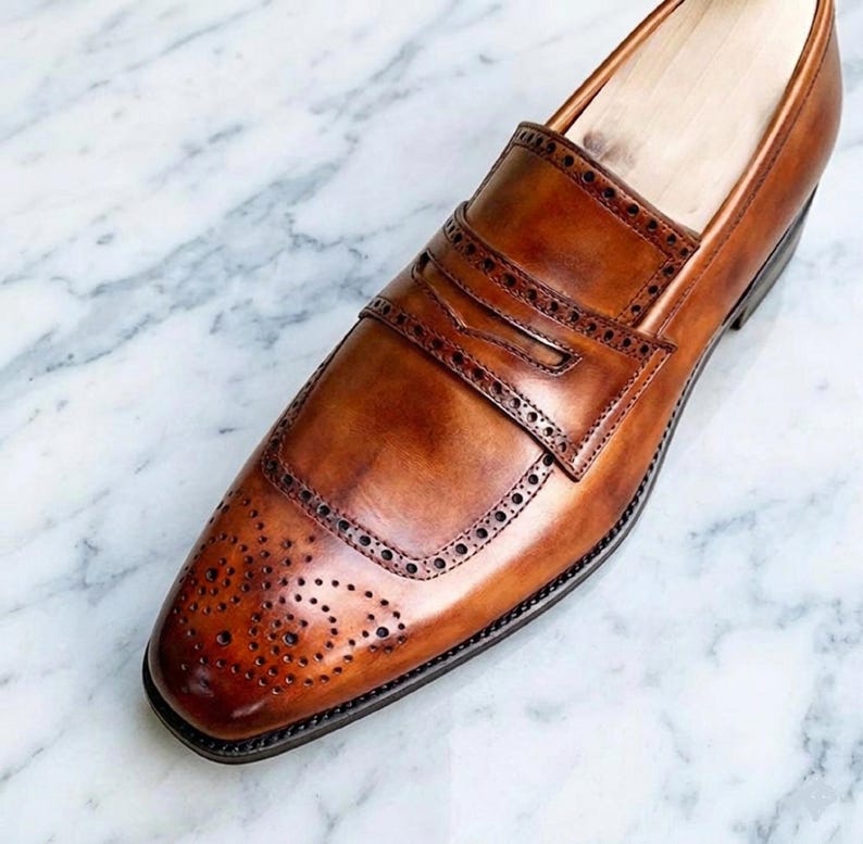 May include: A tan leather penny loafer shoe featuring a burnished finish, a decorative strap across the vamp, and intricate brogue detailing with perforated patterns along the toe cap and the edges of the saddle strap, showcasing a polished look.