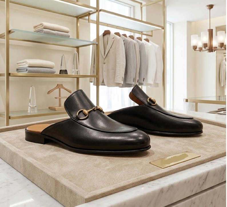 Puede incluir: Mocasines de cuero negro con detalles dorados, expuestos sobre una bandeja beige. Los zapatos tienen un dise&ntilde;o cl&aacute;sico, con punta redondeada y tac&oacute;n bajo. El fondo muestra una tienda de ropa con trajes y estanter&iacute;as.