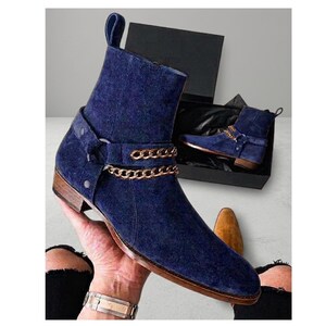 Puede incluir: Botines de ante azul marino con detalles de cadena dorada. Los botines tienen punta afilada y tacón bajo de madera. Un botín se muestra fuera de una caja negra, y el otro dentro.