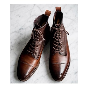 May include: A pair of brown leather lace-up boots featuring a smooth toe cap and a textured leather upper. The boots have dark brown laces, metal eyelets, and a pull tab at the back of the ankle collar. The leather displays a polished finish.