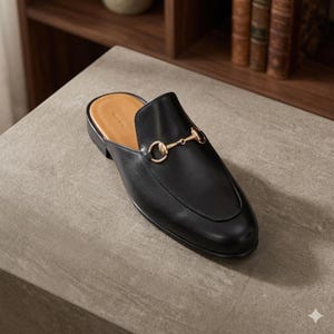 Puede incluir: Mule de cuero negro con un detalle met&aacute;lico dorado. El zapato tiene una punta redondeada y un tac&oacute;n bajo. El interior es de color beige claro. El zapato est&aacute; sobre una superficie gris.