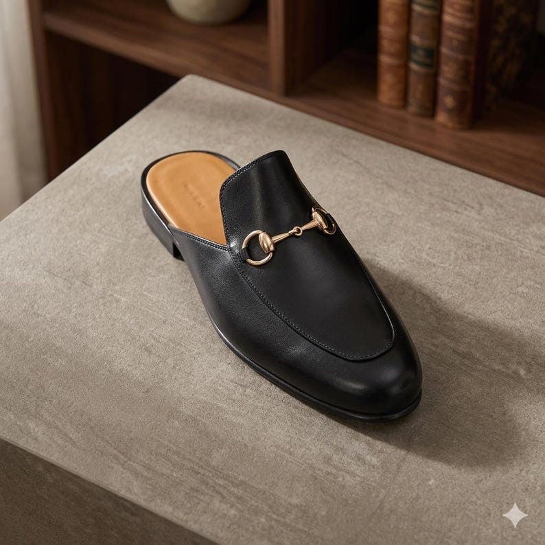 Puede incluir: Mule de cuero negro con un detalle met&aacute;lico dorado. El zapato tiene una punta redondeada y un tac&oacute;n bajo. El interior es de color beige claro. El zapato est&aacute; sobre una superficie gris.
