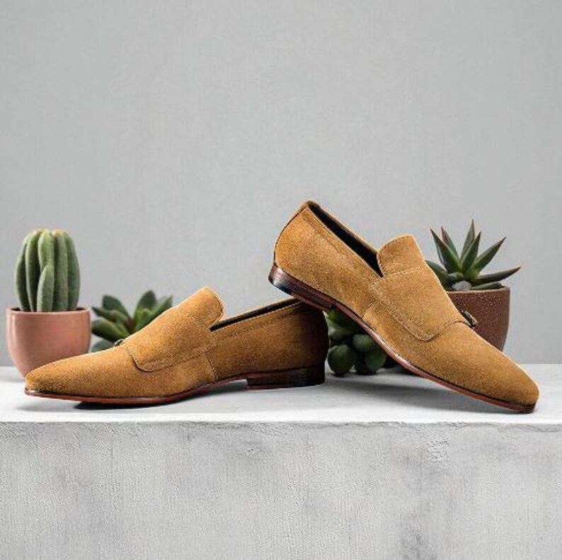 Puede incluir: Un par de mocasines de ante color canela con punta afilada y detalle de correa. Los zapatos se exhiben sobre una superficie blanca con peque&ntilde;as plantas en macetas en el fondo. Suela de cuero.