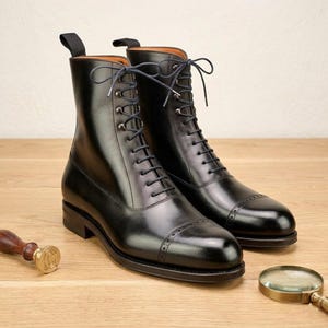 May include: A pair of black leather lace-up boots with a cap toe design. The boots have a high-top silhouette, a pull tab at the back, and are displayed on a wooden surface. The boots are new and unworn.