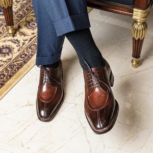 Peut inclure: Une paire de chaussures de ville en cuir marron avec un bout rapporté. Les chaussures ont une finition polie et sont lacées. L'image montre les chaussures d'un angle bas, soulignant leur forme et leurs détails. Les chaussures sont portées avec un pantalon bleu marine et des chaussettes sombres.