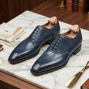 May include: A pair of blue leather Oxford shoes with brogue detailing and black laces. The shoes have wooden shoe trees inside. A gold-coloured shoe horn and a folded handkerchief are also visible.