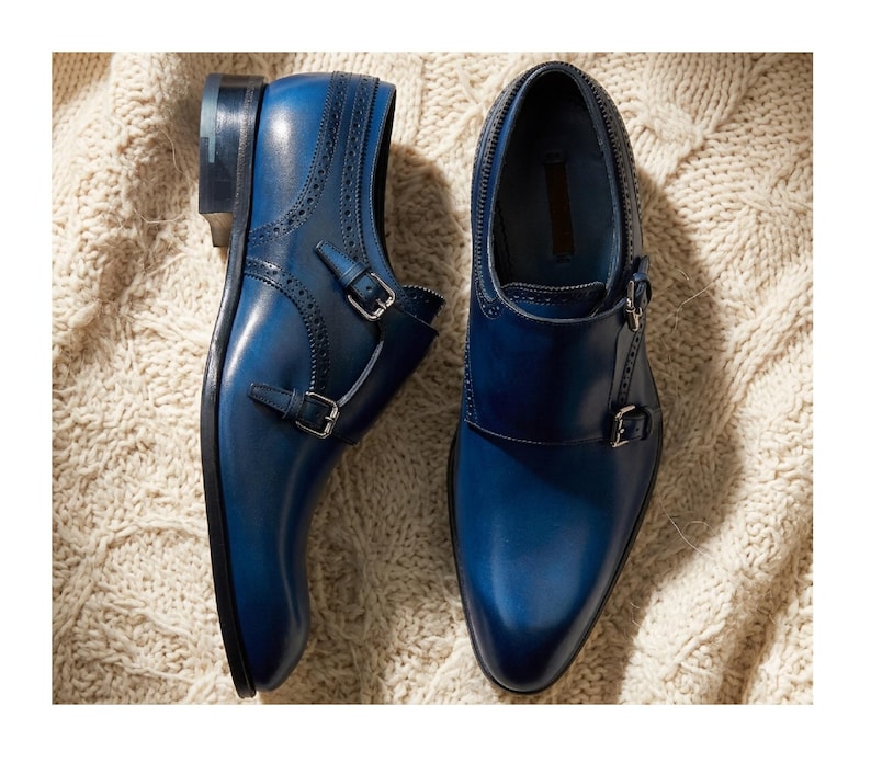 May include: A pair of blue leather double monk strap shoes. The shoes have a pointed toe, decorative perforations, and silver-colored buckles. The shoes are displayed on a cream-colored knitted surface.