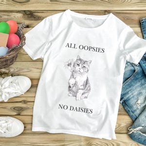 Könnte beinhalten: Weißes T-Shirt mit einer Illustration einer Katze, die eine Blume hält, und dem Text "ALL OOPSIES NO DAISIES". Das Bild zeigt weiße Turnschuhe, einen Korb mit Eiern und eine blaue Jeans auf einer Holzoberfläche.