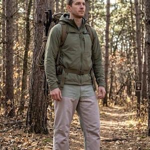 May include: A person wearing outdoor apparel stands in a forest. They are wearing a green jacket, light-colored pants, and hiking boots. A backpack with trekking poles and rope is visible. The background features trees and a path.