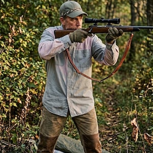 May include: A person in a forest setting aiming a rifle with a scope. They wear a green cap, a light blue and pink button-down shirt, brown gloves, and brown pants with suede patches. A brown leather strap is attached to the rifle.