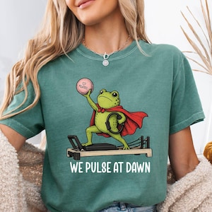 May include: Teal green t-shirt featuring a cartoon frog superhero on a Pilates reformer machine. The frog is wearing a red cape and holding a pink ball. The text "WE PULSE AT DAWN" is printed below the frog. The shirt is paired with denim shorts.