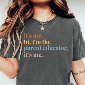 May include: A dark gray t-shirt with the text "it's me, hi. i'm the parent educator, it's me." printed in colorful letters.