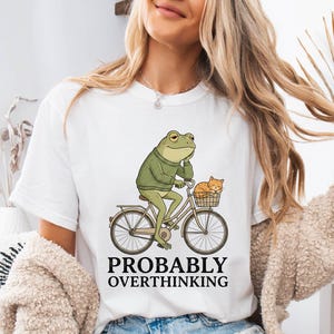 May include: White t-shirt featuring a cartoon frog riding a bicycle with a cat in the basket. The frog wears a green sweater. The text "PROBABLY OVERTHINKING" is printed below the image in black.