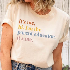 Pu&ograve; includere: Una maglietta color crema con la scritta "it's me, hi. I'm the parent educator, it's me." in lettere colorate. La maglietta &egrave; abbinata a jeans di denim blu chiaro con dettagli strappati.