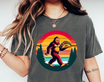 Może przedstawiać: Szary T-shirt z grafiką w stylu retro przedstawiającą Bigfoota trzymającego taco. Grafika umieszczona jest na tle zachodu słońca z drzewami.