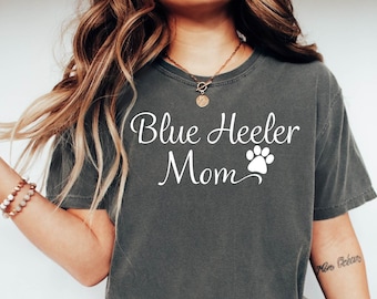 May include: Dark gray t-shirt with the text "Blue Heeler Mom" and a paw print graphic. The shirt is made of a soft material and is paired with blue jeans and white sneakers. A casual outfit for dog lovers.