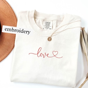 May include: Cream-colored t-shirt with the word "love" embroidered in red script, featuring a heart symbol. A gold necklace with a pendant rests on the shirt. The word "embroidery" is on a white label. A brown hat is visible.