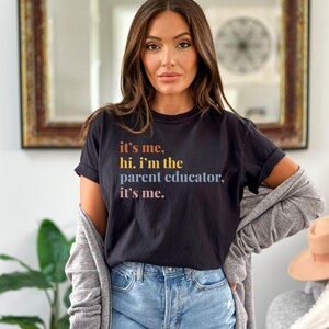 Pu&ograve; includere: T-shirt nero con la scritta "it's me, hi. I'm the parent educator, it's me." in lettere arancioni, gialle e viola. La maglietta &egrave; abbinata a jeans blu e un cardigan grigio.