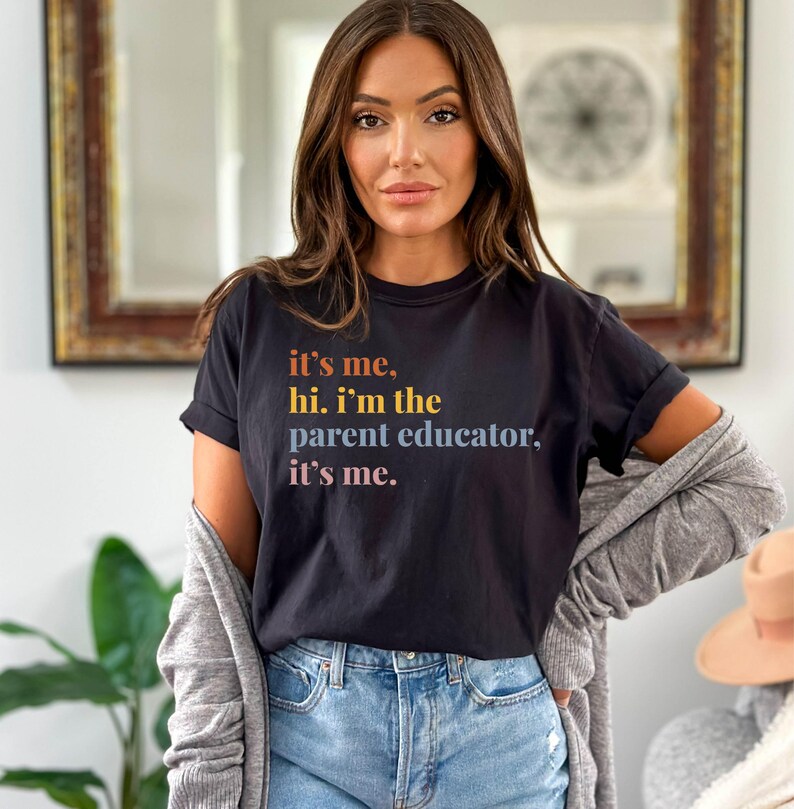 Pu&ograve; includere: T-shirt nero con la scritta "it's me, hi. I'm the parent educator, it's me." in lettere arancioni, gialle e viola. La maglietta &egrave; abbinata a jeans blu e un cardigan grigio.