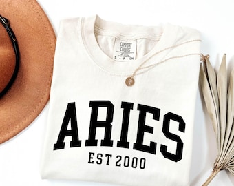 Op de afbeelding: Crèmekleurig T-shirt met het woord "ARIES" in vette zwarte letters en "EST 2000" eronder. Een gouden ketting met een hanger en een bruine hoed staan ook op de afbeelding. Het shirt is van het merk Comfort Colors.