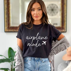 Op de afbeelding: Een zwart T-shirt met witte tekst die "vliegtuigmodus" zegt en een kleine vliegtuigafbeelding.