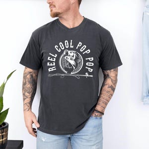 May include: A black t-shirt with a white graphic design of a fish jumping out of the water with the text "Reel Cool Pop Pop" around it.
