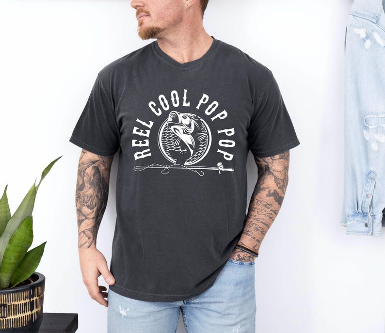 May include: A black t-shirt with a white graphic design of a fish jumping out of the water with the text "Reel Cool Pop Pop" around it.