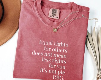 May include: A dusty rose Comfort Colors t-shirt with the text "Equal rights for others does not mean less rights for you. It's not pie. RBG." A silver necklace with a small pendant rests on the shirt.