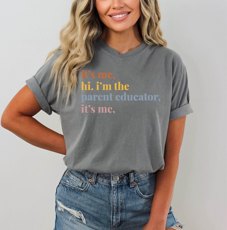 Pu&ograve; includere: T-shirt grigio con maniche arrotolate e la scritta "It's me, hi. I'm the parent educator, it's me." in arancione, giallo, blu e rosa. La maglietta &egrave; abbinata a pantaloncini di jeans.