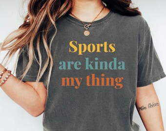 May include: Dark gray t-shirt with the text "Sports are kinda my thing" in yellow, teal, and orange. The shirt has a relaxed fit and short sleeves. A casual, comfortable tee for sports enthusiasts.