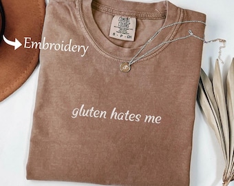 May include: A brown Comfort Colors t-shirt with the embroidered text "gluten hates me." A silver necklace with a small pendant rests on the shirt. A brown hat is partially visible in the upper left corner.