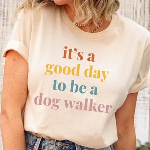 May include: Cream-colored t-shirt with the phrase "it's a good day to be a dog walker" in orange, yellow, teal, and pink lettering. The shirt has rolled-up sleeves and is made of a soft material.