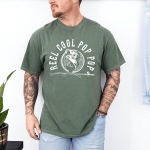May include: A green t-shirt featuring a white graphic of a fish being reeled in with a fishing rod. The graphic is surrounded by the text "Reel Cool Pop Pop".