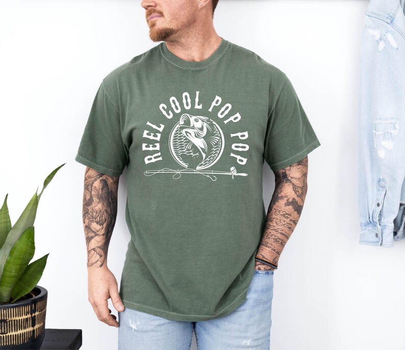 May include: A green t-shirt featuring a white graphic of a fish being reeled in with a fishing rod. The graphic is surrounded by the text "Reel Cool Pop Pop".