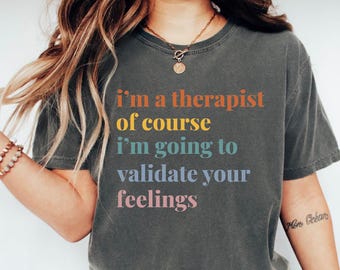 May include: Dark gray t-shirt with the text "I'm a therapist of course I'm going to validate your feelings" in orange, yellow, teal, and pink. The shirt is a casual style.