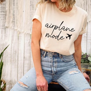 Op de afbeelding: Een wit T-shirt met de tekst "airplane mode" in zwarte letters met een kleine vliegtuigafbeelding. Het model draagt een blauwe jeans.