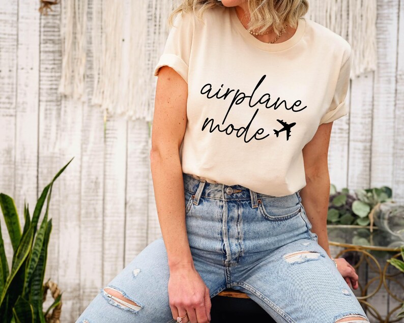 Op de afbeelding: Een wit T-shirt met de tekst "airplane mode" in zwarte letters met een kleine vliegtuigafbeelding. Het model draagt een blauwe jeans.